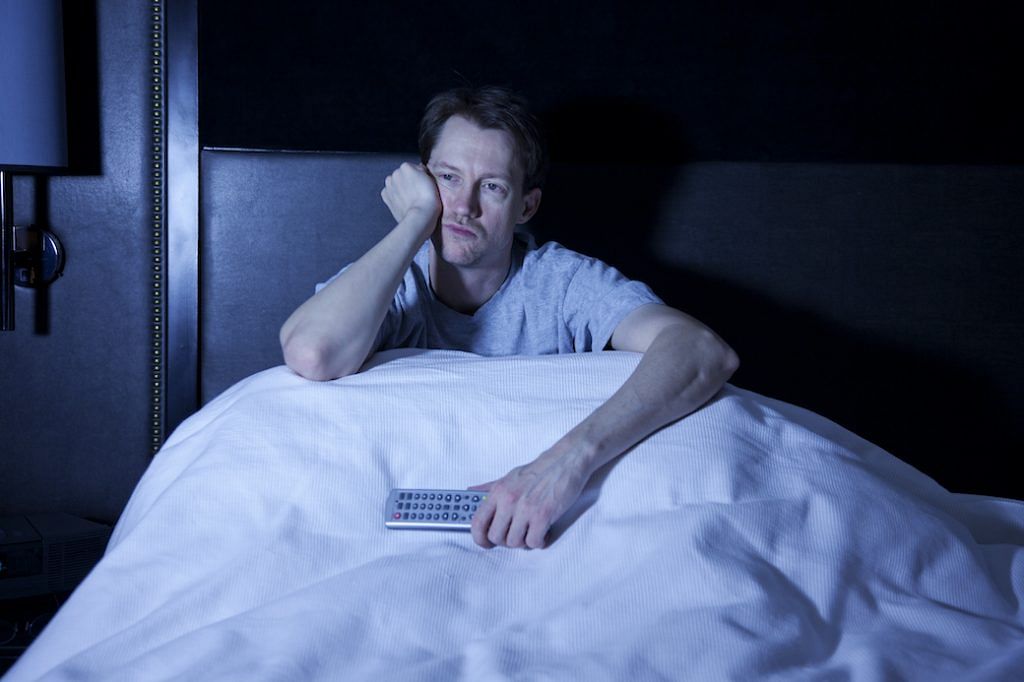 Photo of a man sitting in his bed, watching late night television with remote control in hand.